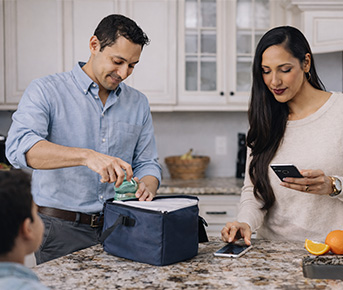 Family morning routine in a bright kitchen—packing a lunch bag while checking a phone and sipping coffee