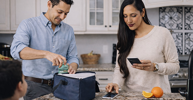 Family morning routine in a bright kitchen—packing a lunch bag while checking a phone and sipping coffee